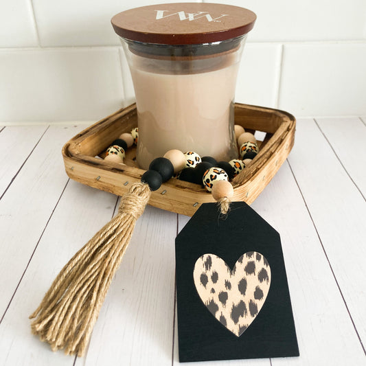 Handmade Leopard print wood bead garland with a variety of black natural and leopard printed beads jute twine tassel and black painted tag with leopard print heart 24 inches in total length displayed in a tobacco basket with a candle