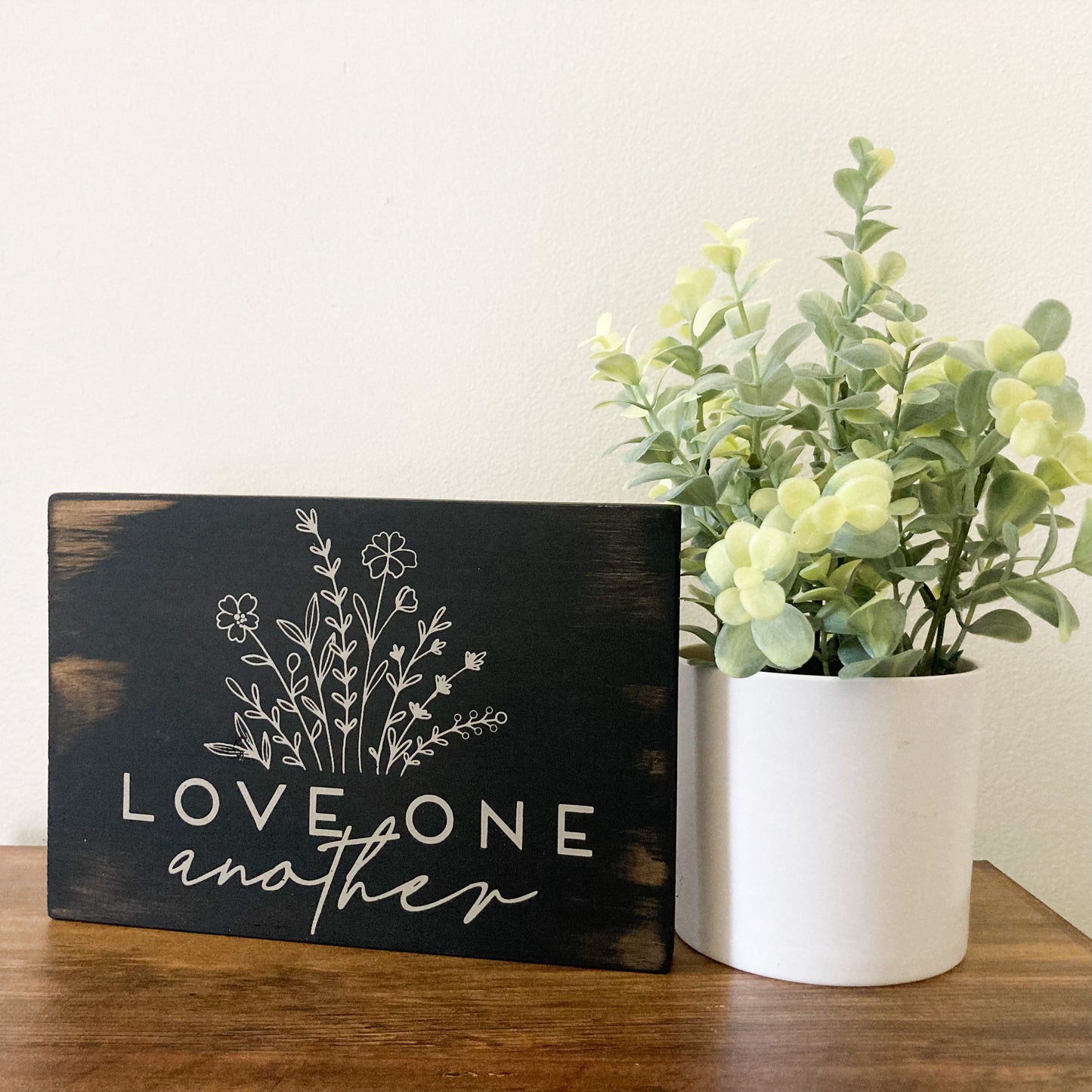 Handmade wood sign painted distressed black with flowers and words saying love one another in white 5.5 inches by 8 inches