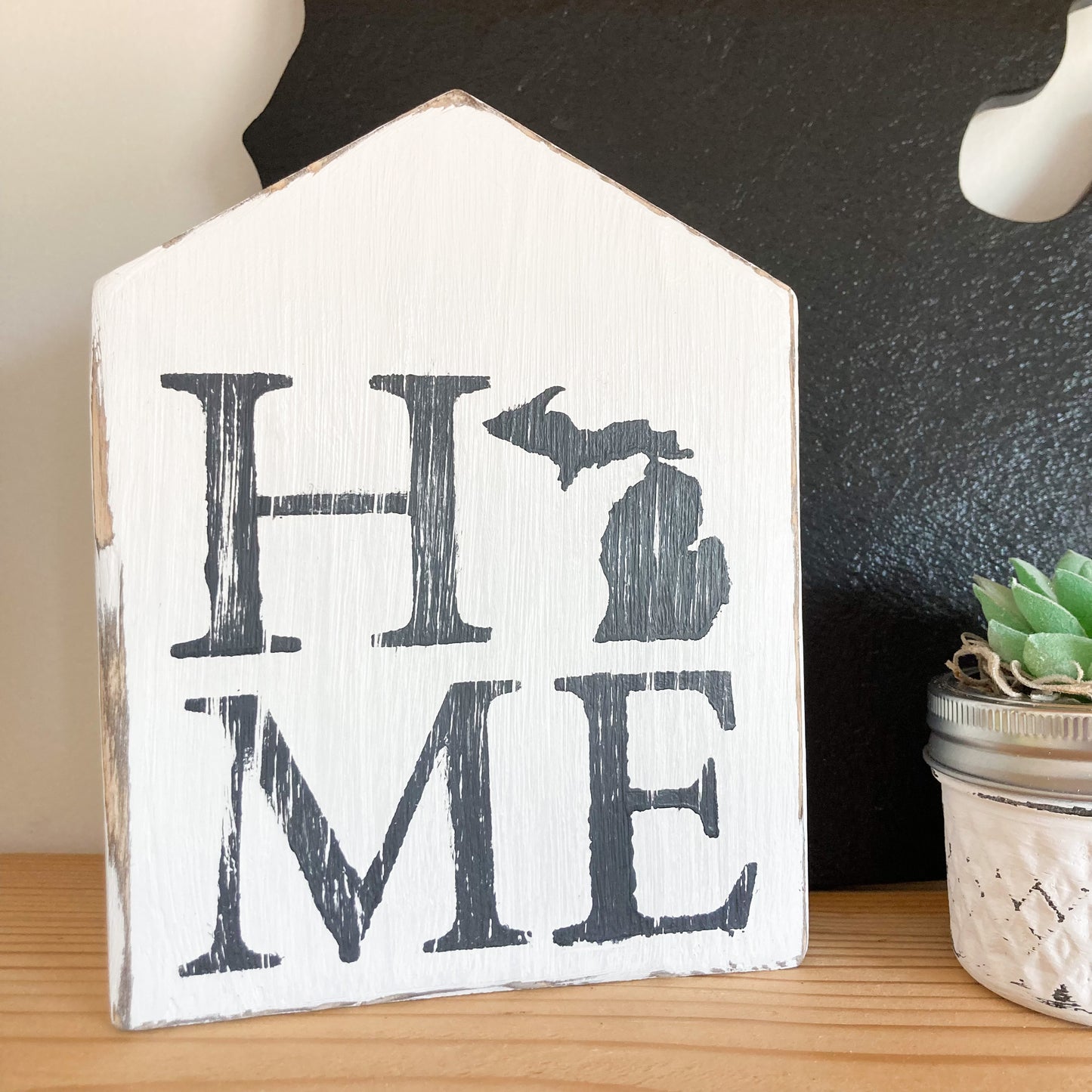 Handmade wood shaped distressed house painted white displaying word home with state of Michigan as the o painted in black approximatly 7 inches by 5 inches by 1.5 inches thick free standing shelf sitter