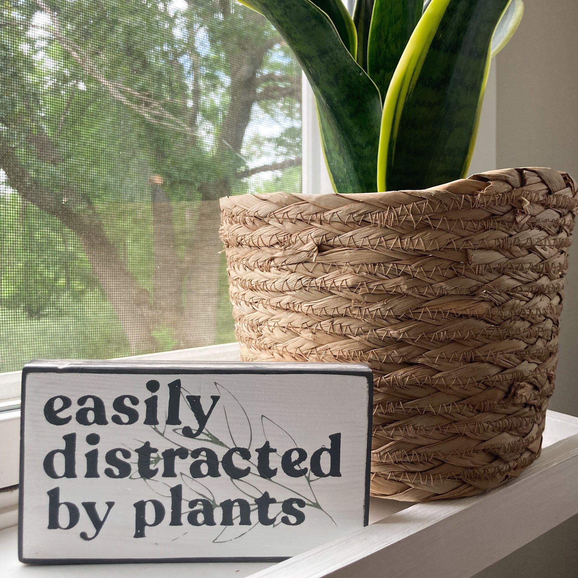 Easily distrated by plants wood sign painted white with black wording and leaf displayed on a window shelf next to a plant 3.5 inches by 6 inches