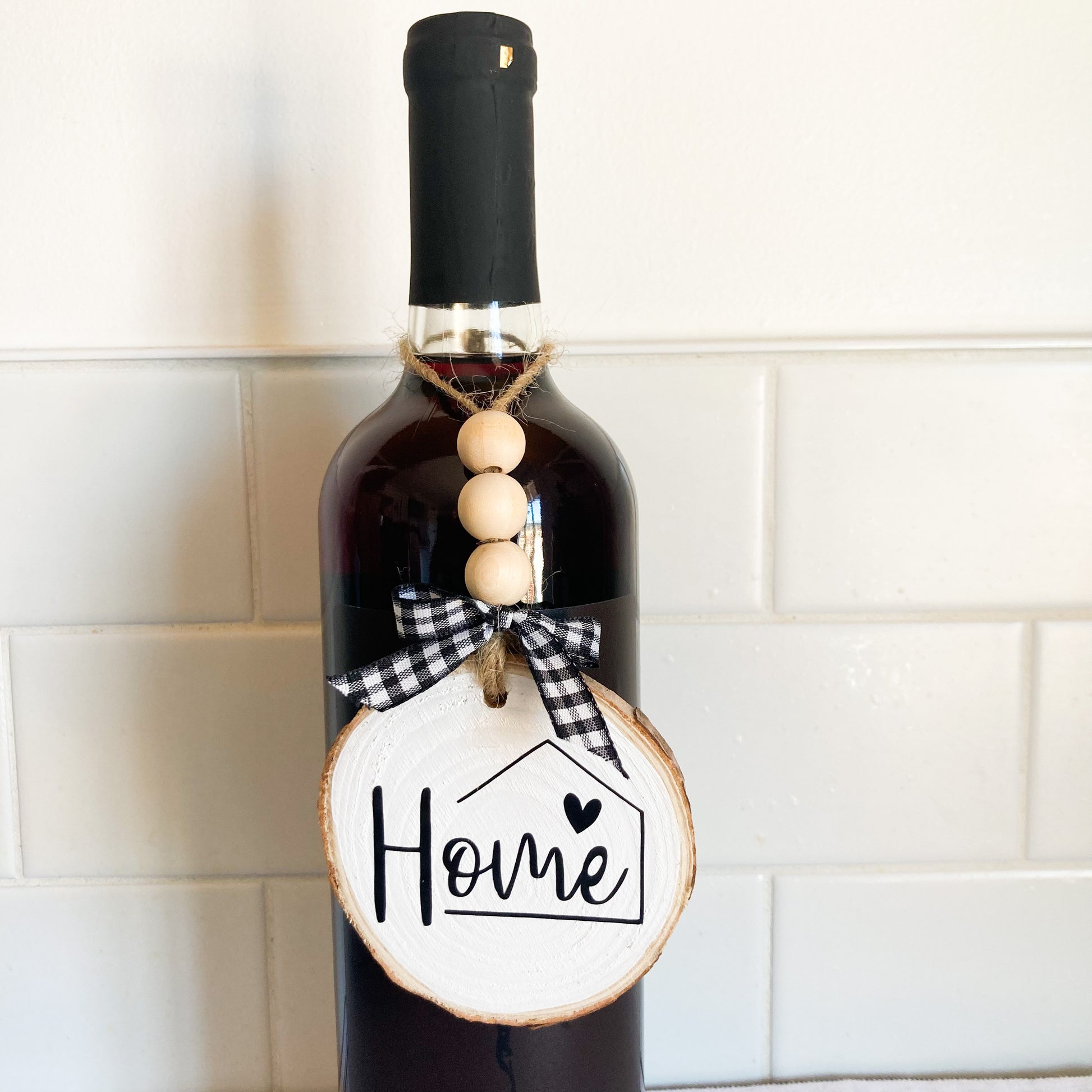 Handmade home ornament on 3 inch wood round painted white with black vinyl word Home jute twine hanger with wood beads and buffalo checkered bow displayed on a wine bottle